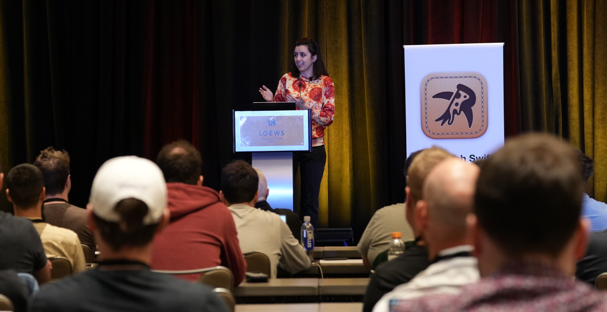 Cassidy speaking at a podium, wearing a pizza shirt